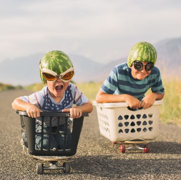 a young boy races his brother in a makeshift go cart while wearing watermelon helmets and goggles he is excited as he is winning the race