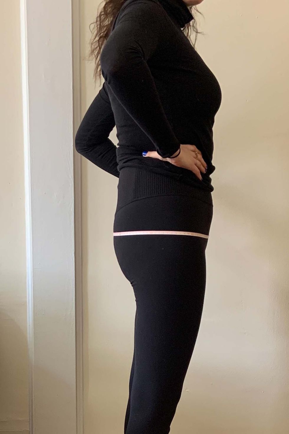 a woman wearing black leggings and a black turtleneck demonstrating how to measure your hips with the measuring tape over the fullest part of her booty