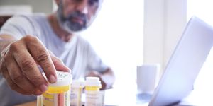 Hispanic Man Sitting At Dining Room Table Reaches For His Prescription Medications