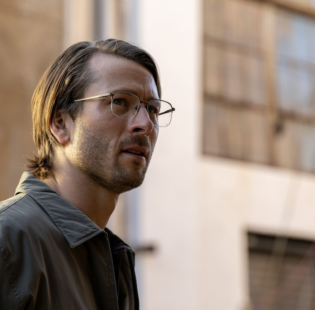 glen powell with long hair and glasses standing in front of an old warehouse in a scene from hit man