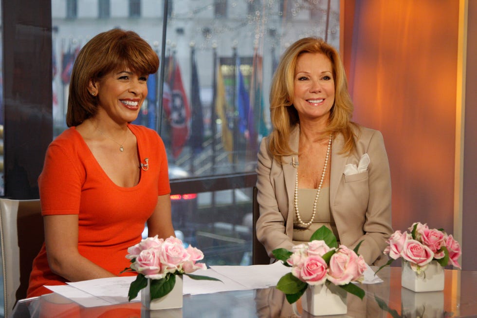 hoda kotb and kathie lee gifford smile while sitting together at a table with multiple rose flower arrangements on it, kotb wears an orange short sleeve dress, gifford wears a tan suit