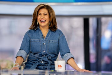 hoda kotb smiles while seated at a table with papers and cups in front of her, she wears a blue jean jumpsuit