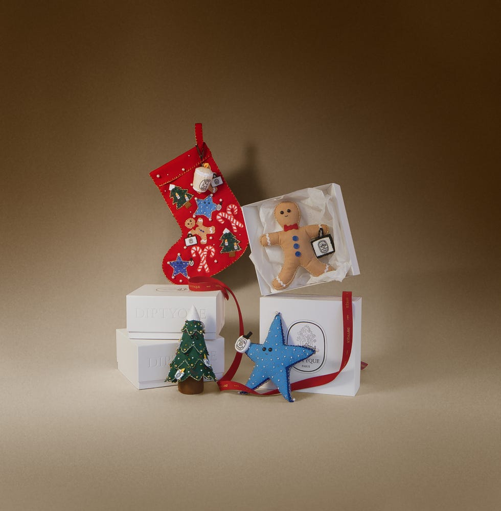 the scene showcases a vibrant red christmas stocking adorned with decorations, positioned next to multiple white boxes with the name 'diptyque' among the items are a gingerbread man ornament, a blue star ornament, and a green christmas tree figure, creating a warm and festive atmosphere