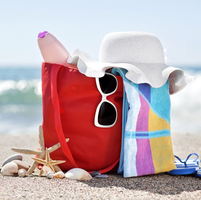 beach bag on the sand filled with sunglasses, a sunhat, sunscreen, and other objects
