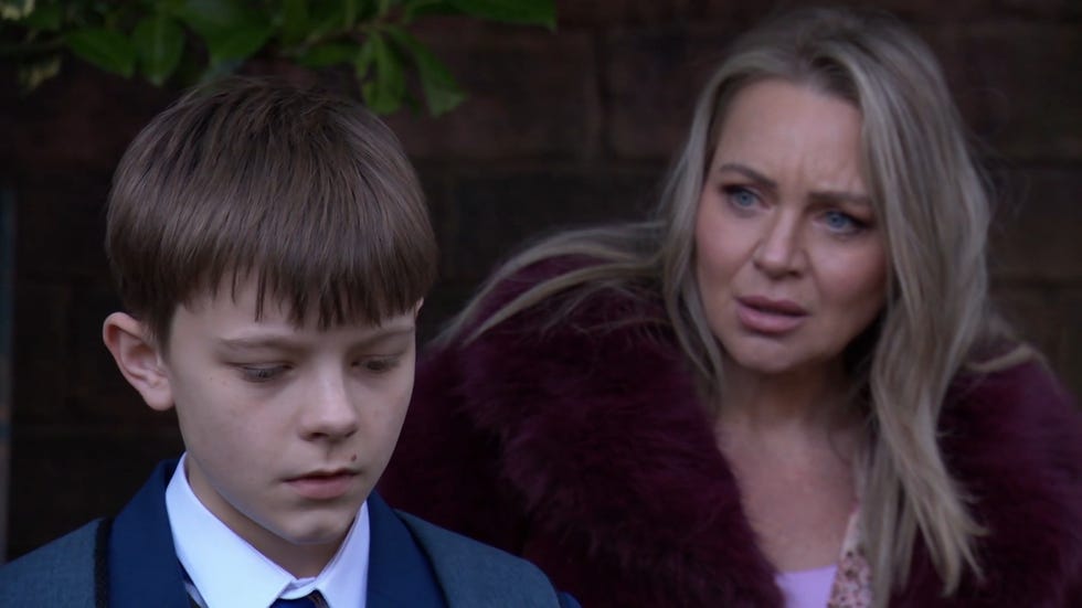 hollyoaks, arlo in a school uniform, with a slightly out of focus marie behind him, wearing a puffy purple coat