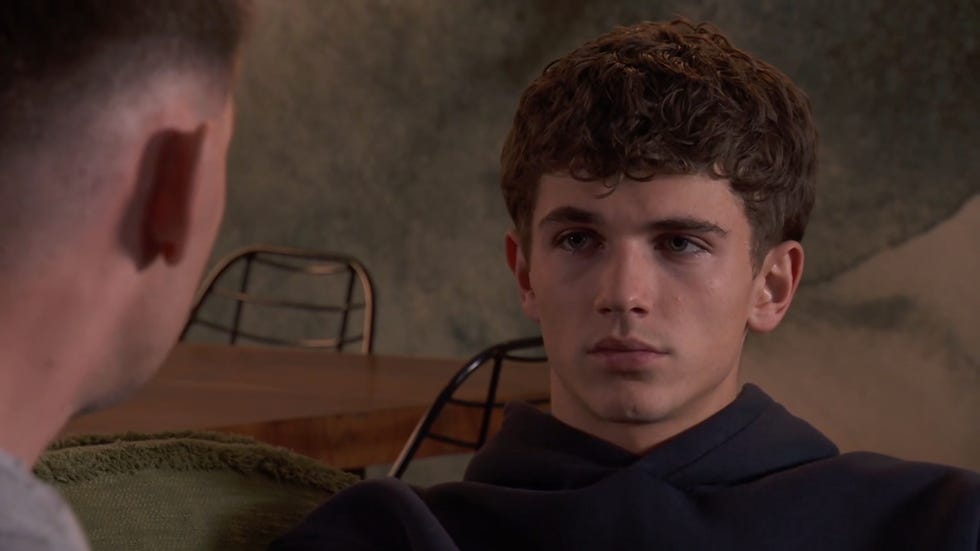 hollyoaks lucas hay looking moody in a dark blue hoodie sitting on a sofa, with ste looking at him