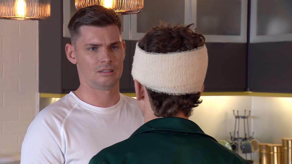 hollyoaks, ste and lucas hay in june 2025. ste in a white t shirt backs off as lucas, facing away from camera in a green hoodie with a bandage around his head, angrily confronts him
