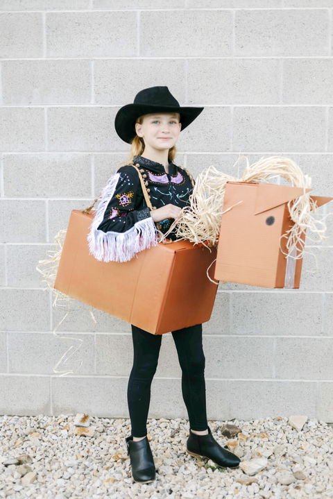 homemade halloween costumes western box costume, girl outside wearing a black cowgirl hat while standing inside a cardboard box shaped like a horse
