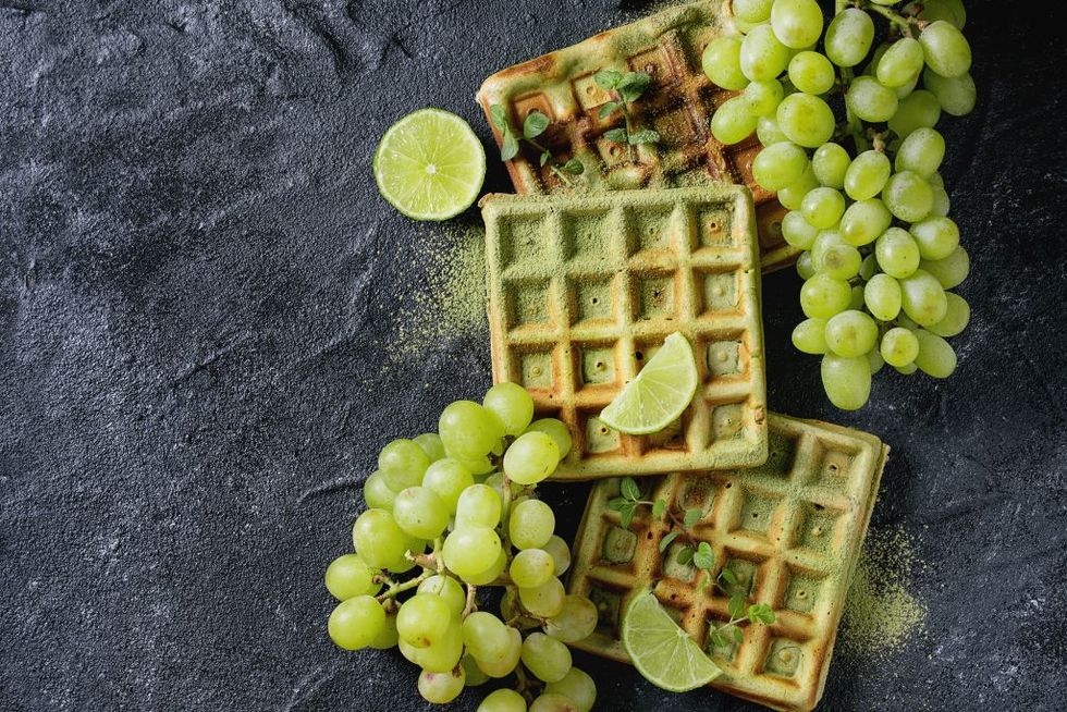 Homemade square green matcha belgian waffles with fresh ripe grapes, mint and lime over black stone texture background. Top view with space