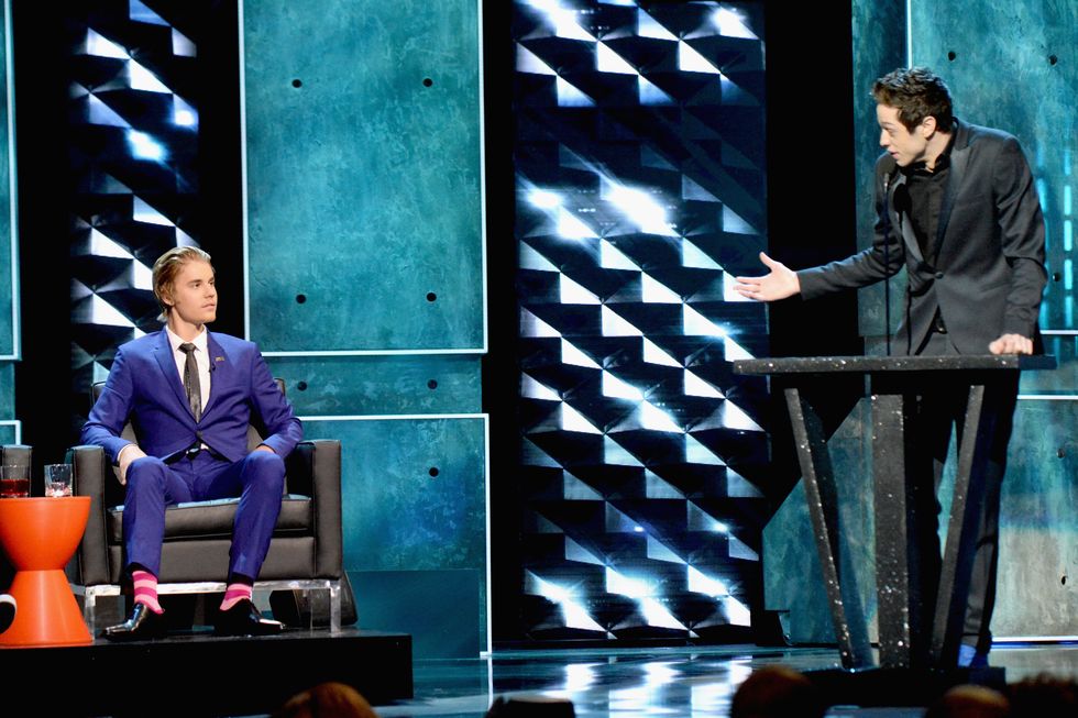 pete davidson, wearing a black suit jacket and black shirt, stands at a podium on a stage and speaks directly to justin bieber, who sits in a black chair on a platform, wearing a blue suit and black tie