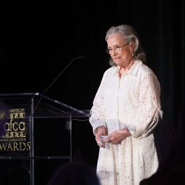 a woman in a white lace dress and glasses stands to the right of a clear podium and smiles, she holds a folded piece of paper in her hands