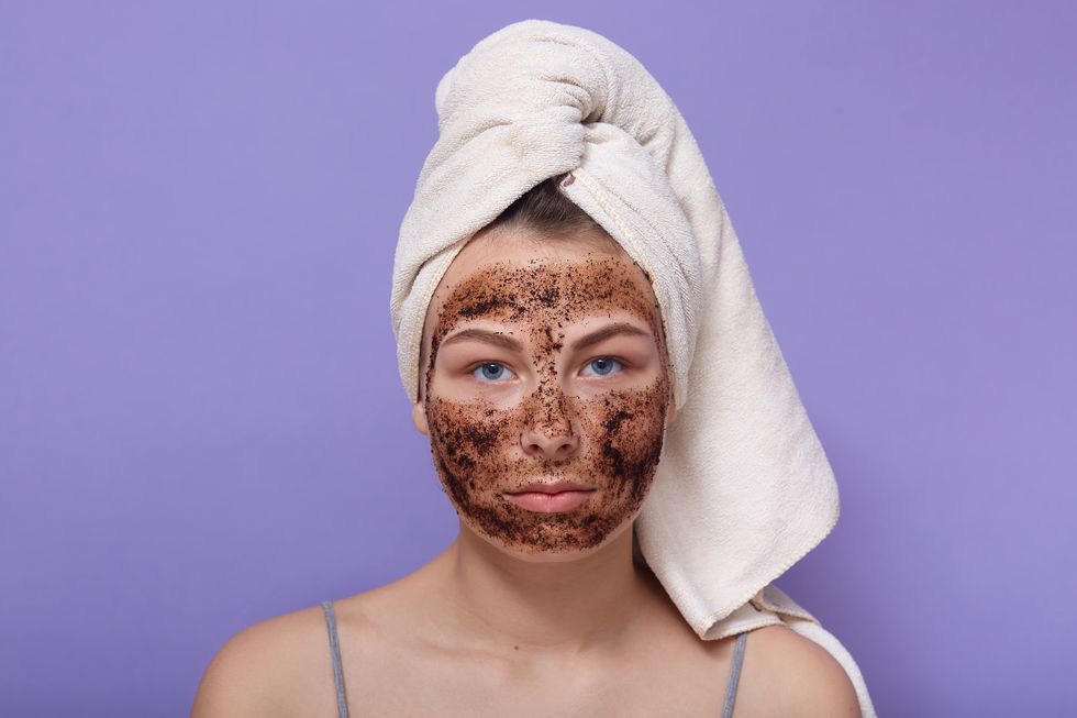 horizontal shot of serious attractive young female looking directly at camera, having applied coffee scrub on face, cleaning skin, having towel on head, home beauty procedures beauty concept