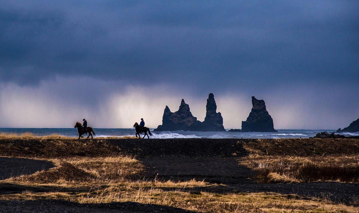 Twee ruiters rijden langs de zuidelijke kust van IJsland en passeren de stacks bij Reynisdrangar