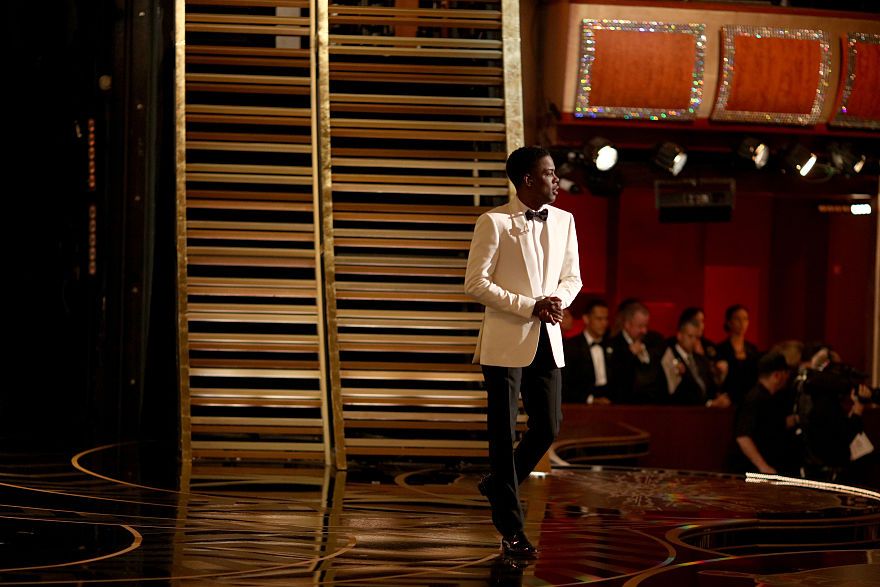 as seen from backstage, chris rock walks around a theater stage, he is looking out toward the audience and his hands are clasped together, audience members are visible in the background, chris is wearing a suit with black pants, a white suit jacket and shirt and a black bow tie