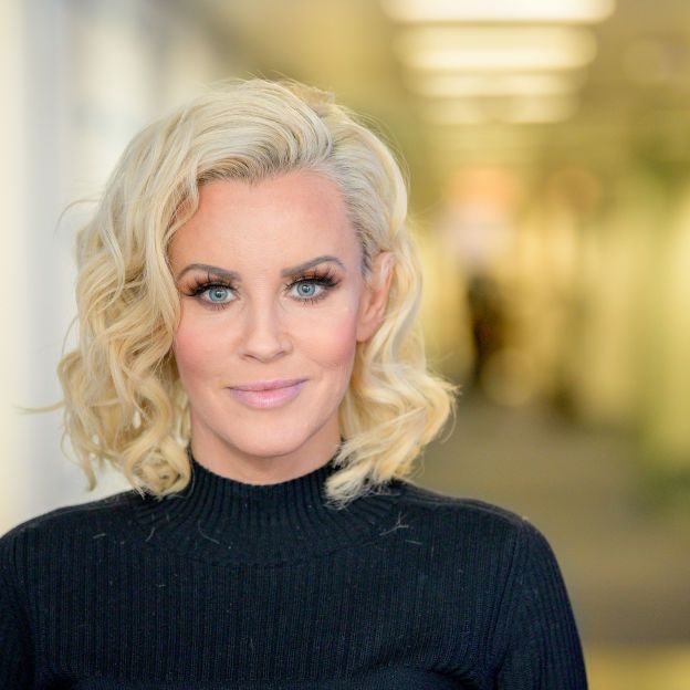 jenny mccarthy looks at the camera with a soft smile during a visit to sirius xm in 2019, she is wearing a black shirt and her blonde hair is curled, touching just at the top of her shoulders