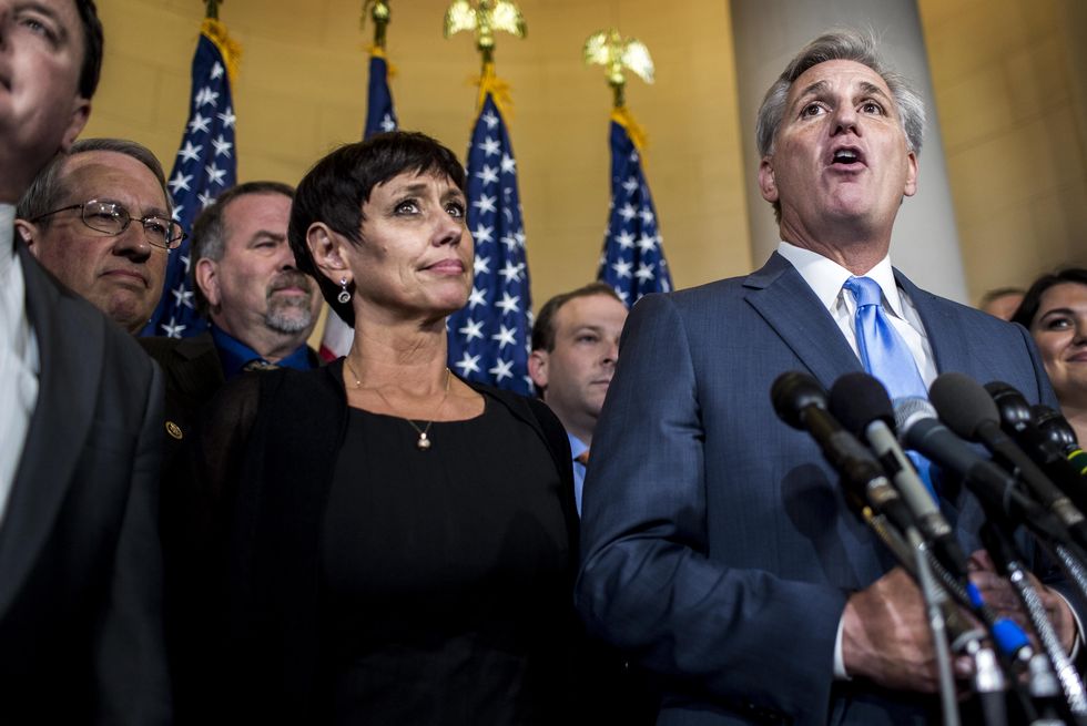 judy mccarthy stands next to kevin mccarthy who is speaking into several microphones, several people surround them, she wears a black outfit and jewelry, he wears a blue suit and tie