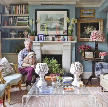 paul hawkins holding his dog in the living room of his colourful london home with blue walls, art work, a stone fireplace and perspex coffee table