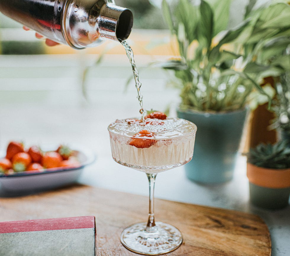 a tall, cut cocktail glass sits on a wooden table beside books, strawberries and plants, as a blended alcoholic drink is poured from a cocktail shaker onto ice and strawberries