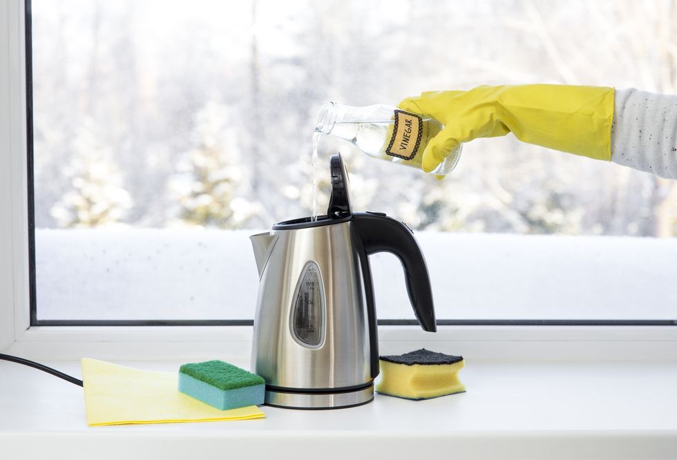 a hand wearing a yellow rubber glove pours white vinegar into an electric kettle to demonstrate how to clean it