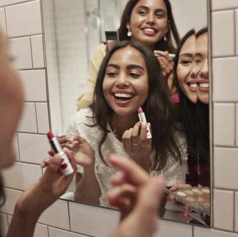 smiling female friends applying lipstick while reflecting in mirror at home