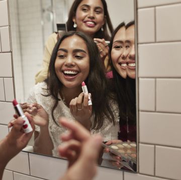 smiling female friends applying lipstick while reflecting in mirror at home