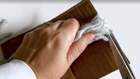 how to make a yarn tassel, woman's hands cutting cardboard yarn