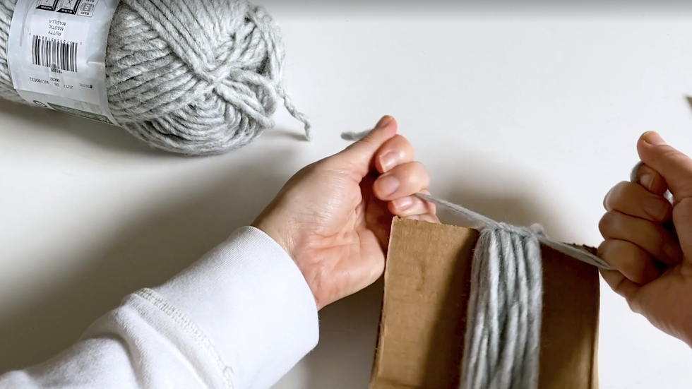 how to make yarn tassel, woman's hands knotting top of gray tassel on cardboard