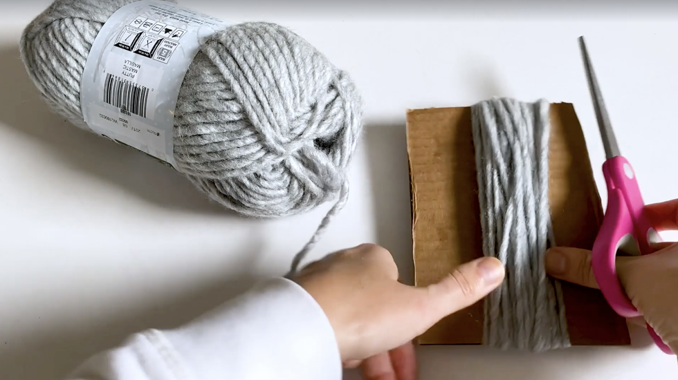 how to make yarn tassel, hand holding pink scissors and cardboard with gray yarn
