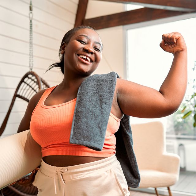 cheerful black woman with a curvy body type in an orange sport bra showing off her bicep while holding a yoga mat and towel