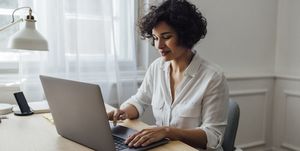 a woman in a white shirt types at her laptop in a home office