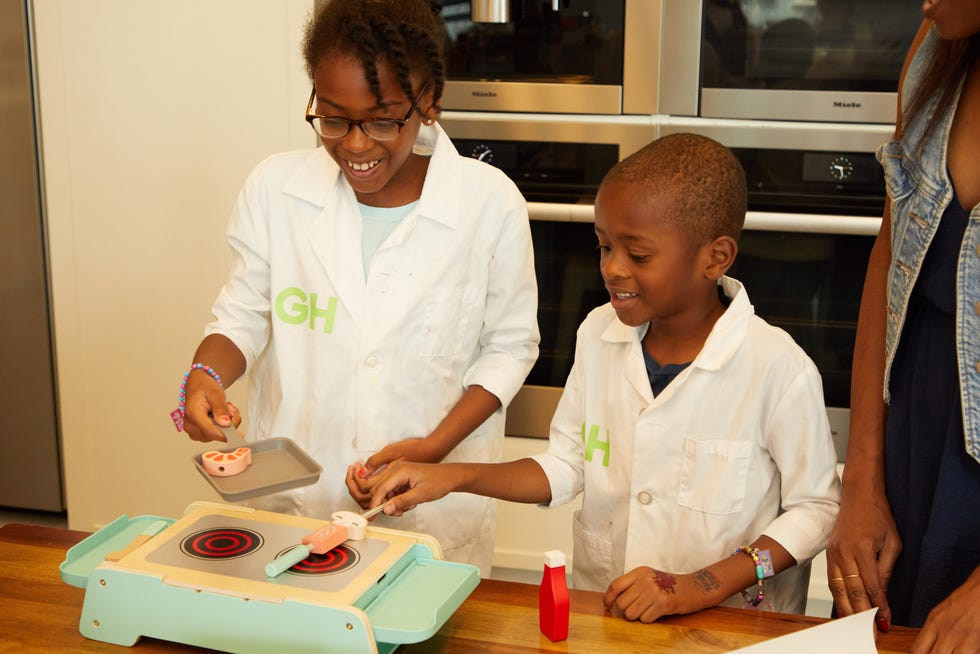 two kids pretend to cook food over a play grill with wooden food pieces they're wearing good housekeeping lab coats