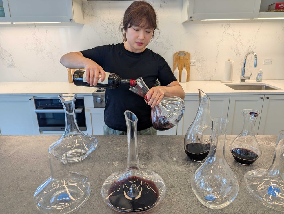 best decanters for wine woman pouring bottle of red wine into a decanter at counter with several other decanters