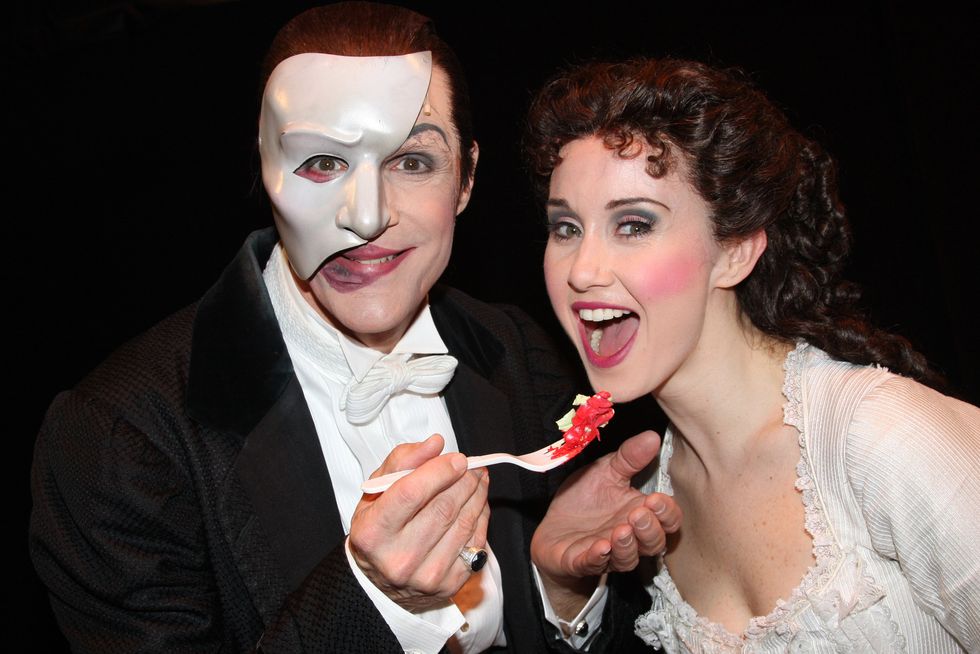 howard mcgillin and elizabeth loyacano dressed as the phantom of the opera characters, with mcgillin holding a plastic spoon with cake up to loyacano's open mouth