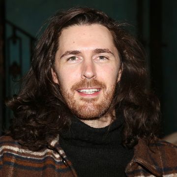 a man with shoulder length curly hair smiles at the camera, he wears a black shirt and a brown white and black plaid jacket