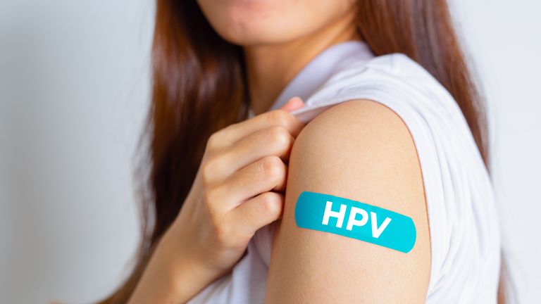 teenage woman showing off an blue bandage after receiving the hpv vaccine, and the bandage reads hpv