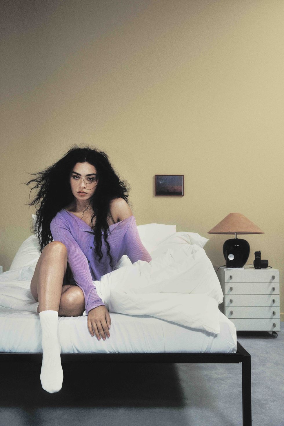 charli xcx sits on the edge of a bed wearing a purple henley