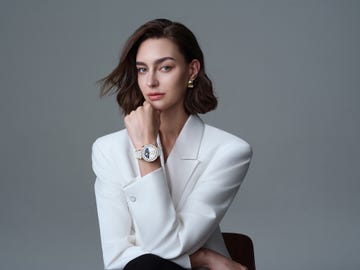 a woman poses with her hand resting on her chin, she is wearing a white blazer and a white and gold smartwatch
