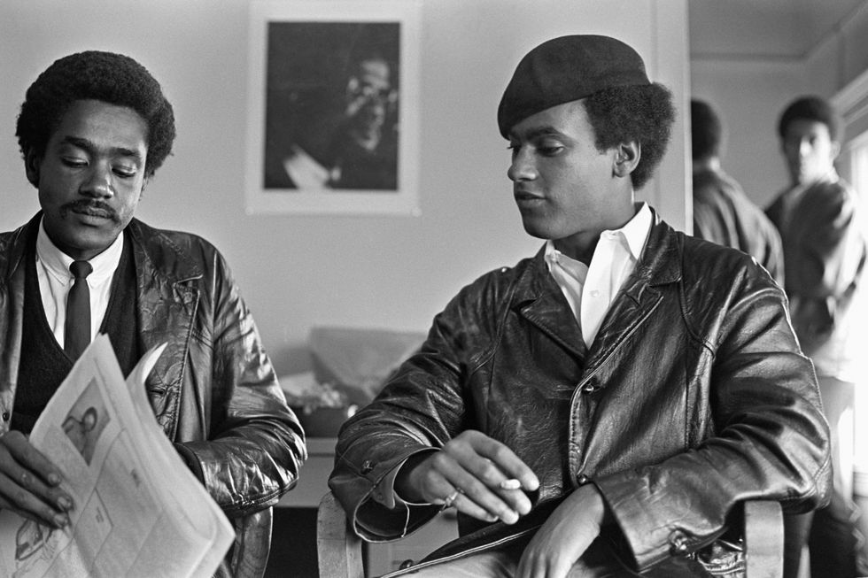 bobby seale and huey p newton sit in chairs next to one another in a living room, both men wear leather jackets and newton has a beret on