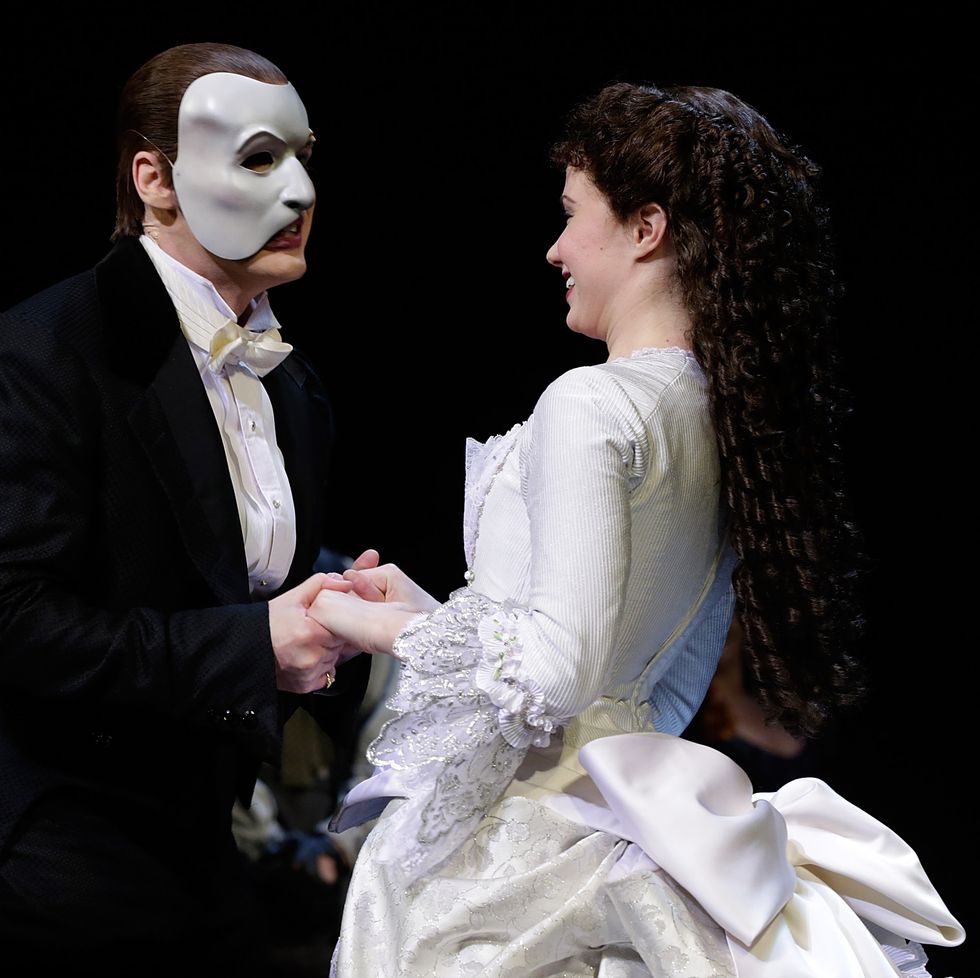 hugh panaro, dressed as the phantom of the opera, holding hands with sierra boggess, wearing a white dress, on a darkened stage