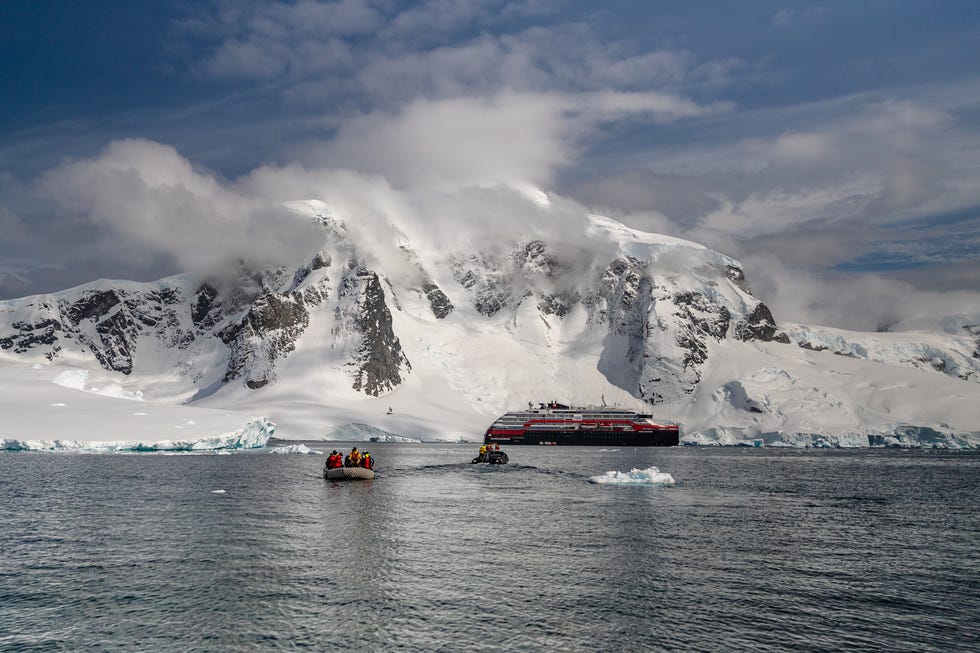 a boat carrying passengers approaches a large ship amidst snowy mountains