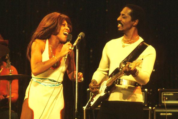 tina and ike turner perform onstage, tina is singing into a handheld microphone and looking at ike who is playing guitar and looking back at her