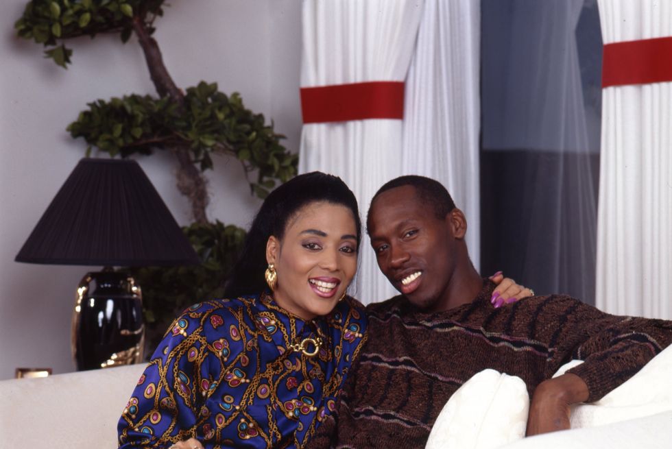 florence joyner and al joyner sit on a couch together and smile at the camera, she wears a blue and gold patterned long sleeve blouse with black pants and bright pink nails, he wears a brown striped sweater and dark khakis, behind them is a window with drapes, a lamp and an indoor plant