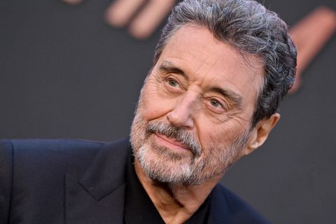 actor ian mcshane, wearing a black suit jacket and black shirt, smiling and looking off camera