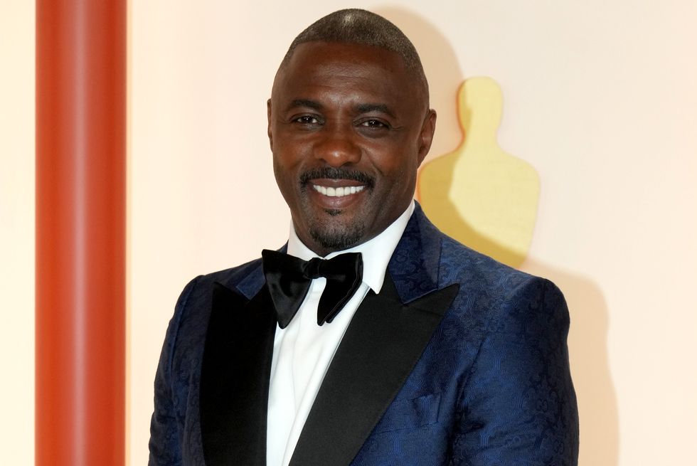 idris elba stands with hands clasped in front and smiling at the camera, closely shaved head with dark hair, wearing a blue tuxedo jacket with black trousers and bow tie and white shirt
