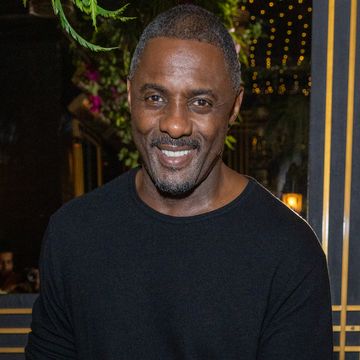 idris elba smiles at the camera, he wears a black shirt and flowers and lights are hanging from the ceiling behind him
