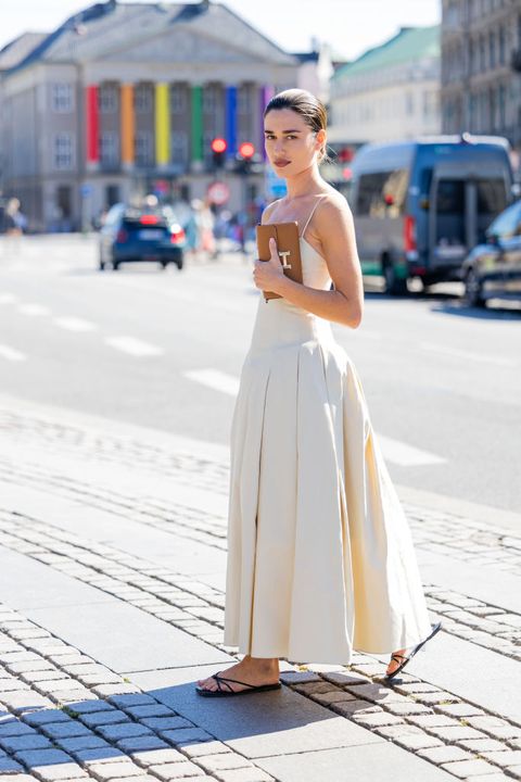 a woman wearing a cream drop waist maxi dress with flat black sandals and a brown hermes clutch on the street during copenhagen fashion week in a roundup of cute summer outfits 2023