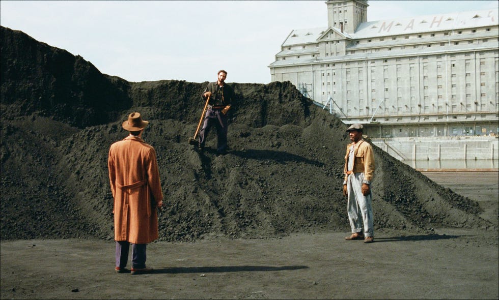 three men engaged in a conversation near a large pile of black material with a large building in the background