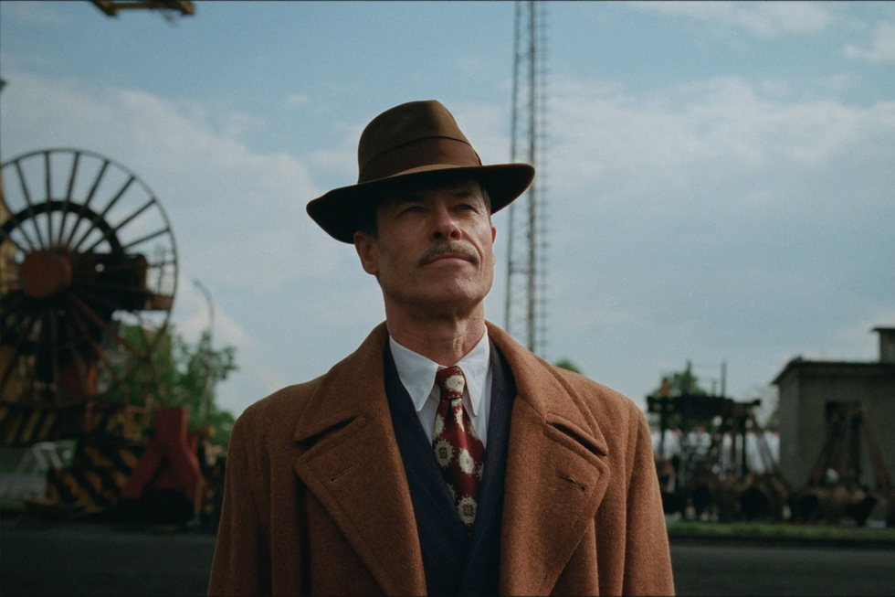 a man in a brown overcoat and hat stands outdoors with industrial machinery in the background