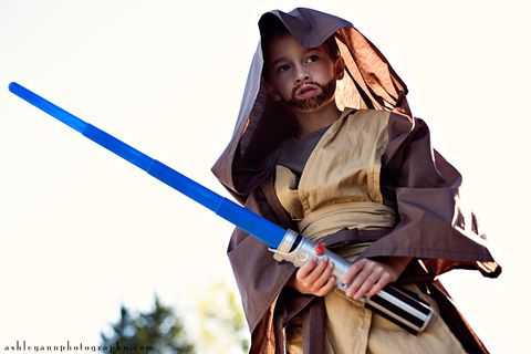 little boy dressed as obi wan kenobi in brown robes with hood, beard drawn on his face, holding a blue light saber