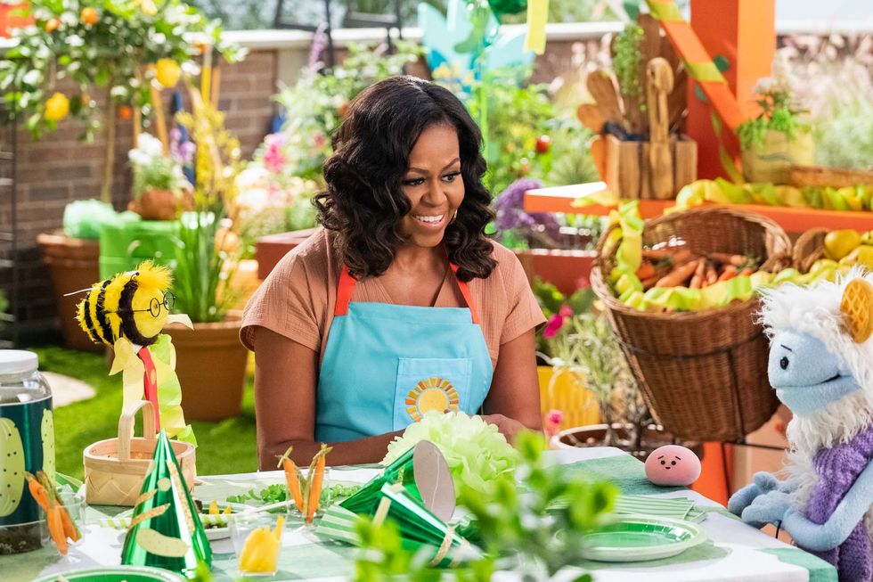 on a rooftop garden, michelle obama, wearing a blue apron, converses with a furry white and blue puppet with frozen waffle ears, a round, pink mochi puppet, and a bee puppet wearing a red tie around a table dressed with green decorations for a birthday party
